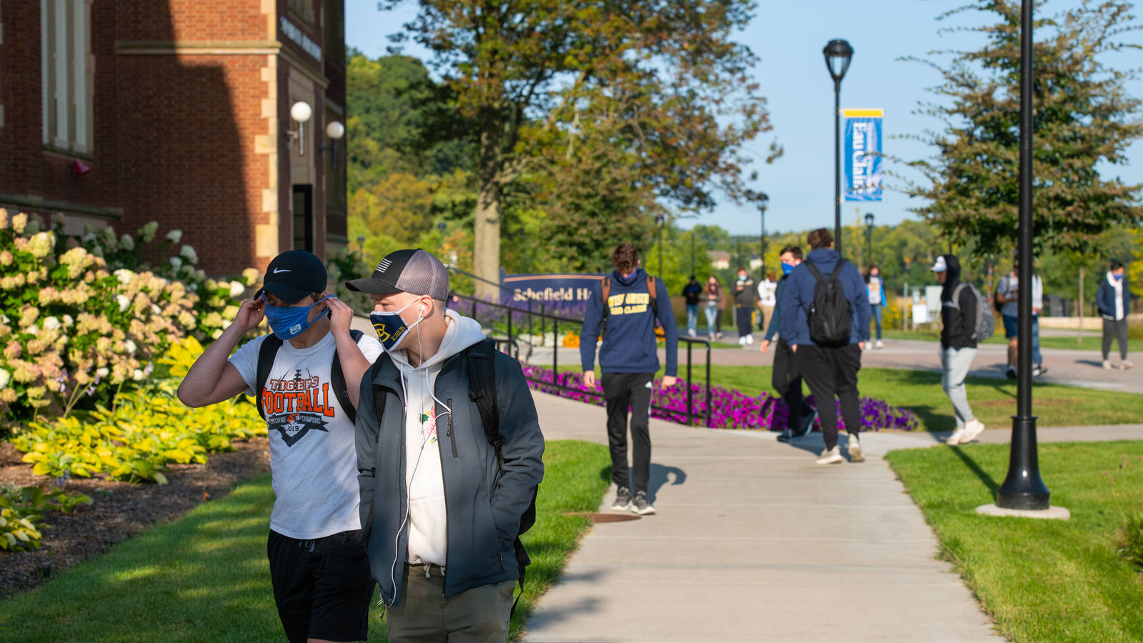 Students walking on sidewalk next to Schofield Hall, masks on, sunny day