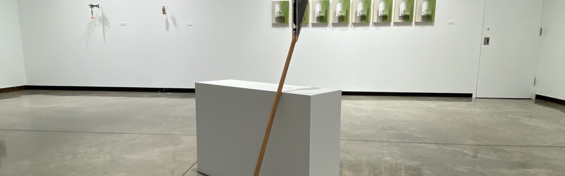 View of "I Make This, You Make That" art exhibition in the Foster Gallery. It shows a sculpture of a broom with a plant growing in a plexi glass container attached to the broom handle.