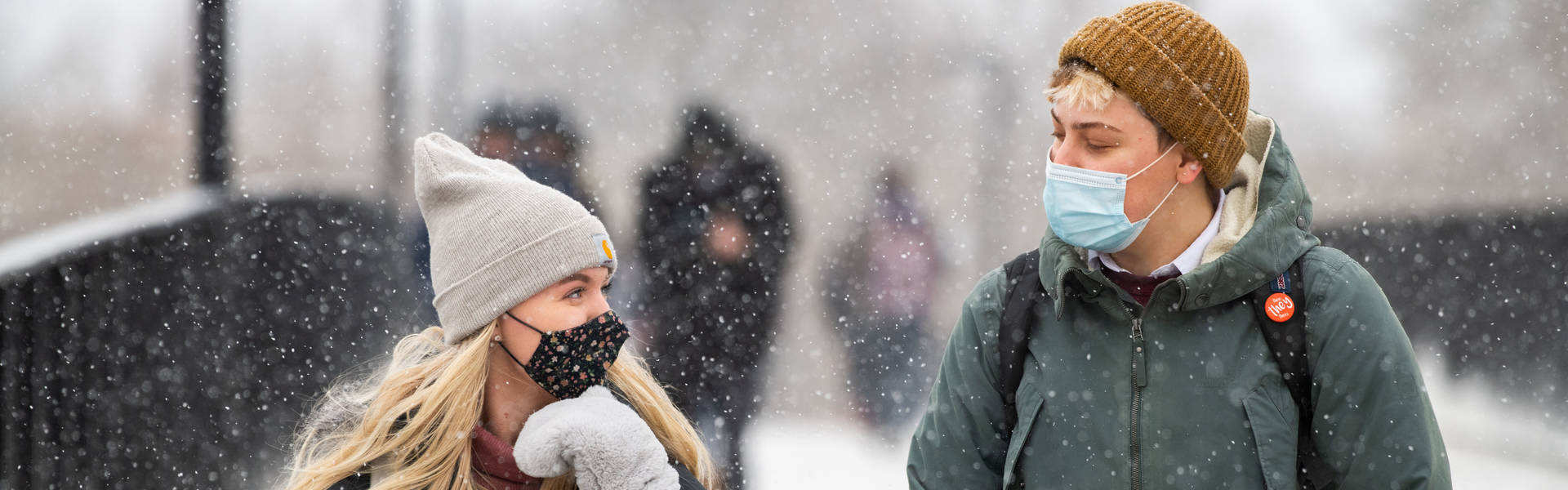 Two students wearing masks smile at each other on the footbridge while it snows.