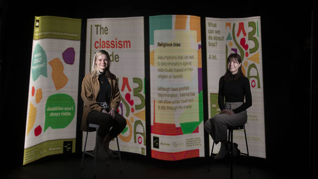 Female students on stools in front of display of their graphic design posters
