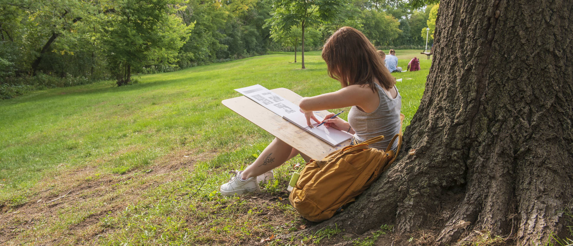 An art student draws on a clipboard next to a tree on campus.