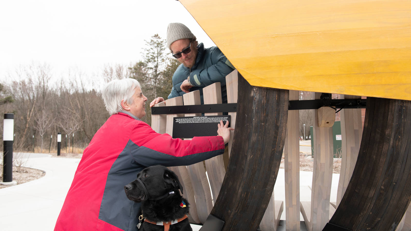 Local artist Joe Maurer and counselor emerita Kathie Schneider with Seeing Eye Dog Calvin explore the Braille plaque installation that features Nick Butler’s poem, Sanctuary, which is also inscribed in the bench’s upper interior.