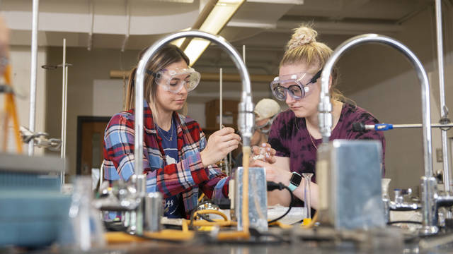 Students wearing goggles conduct an experiment in a chemistry lab.