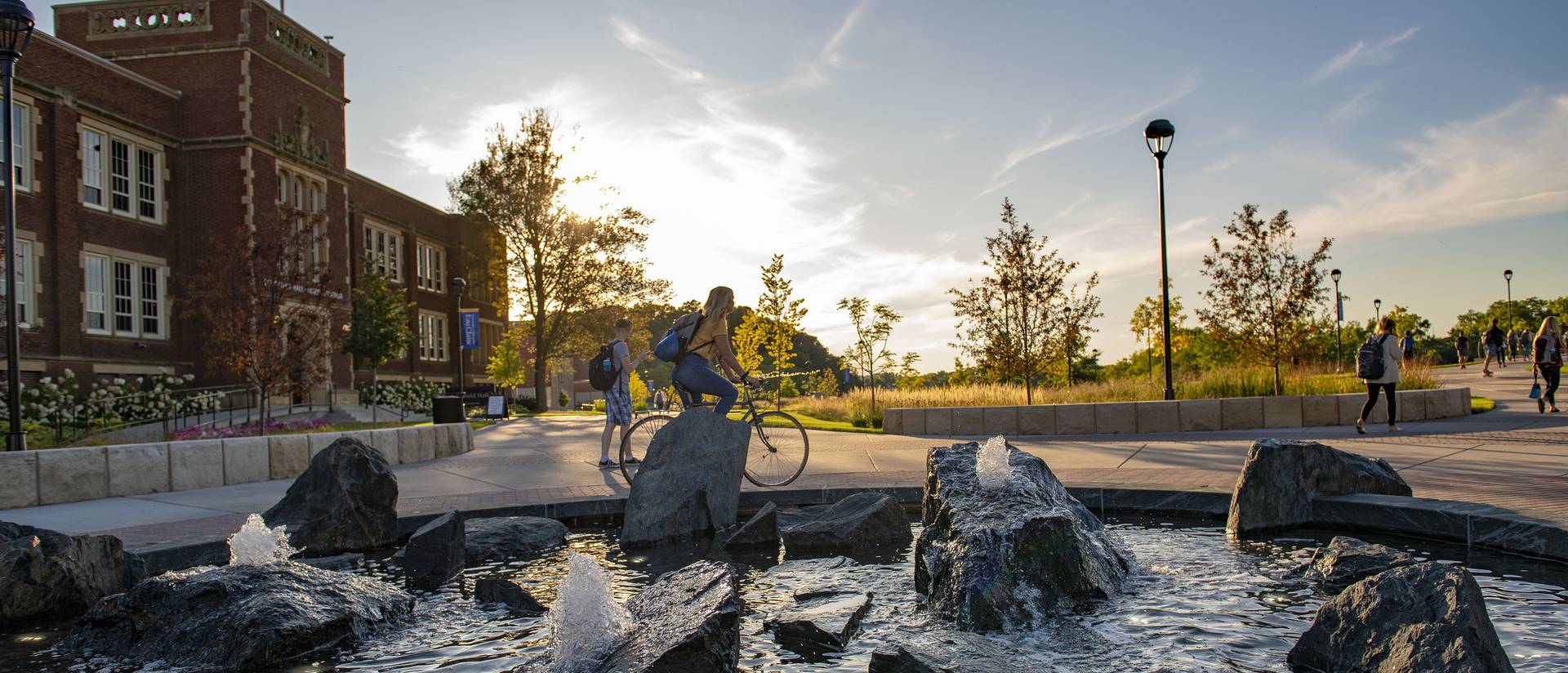Students walk and bike next to the Stowe Fountain during a sunset on campus