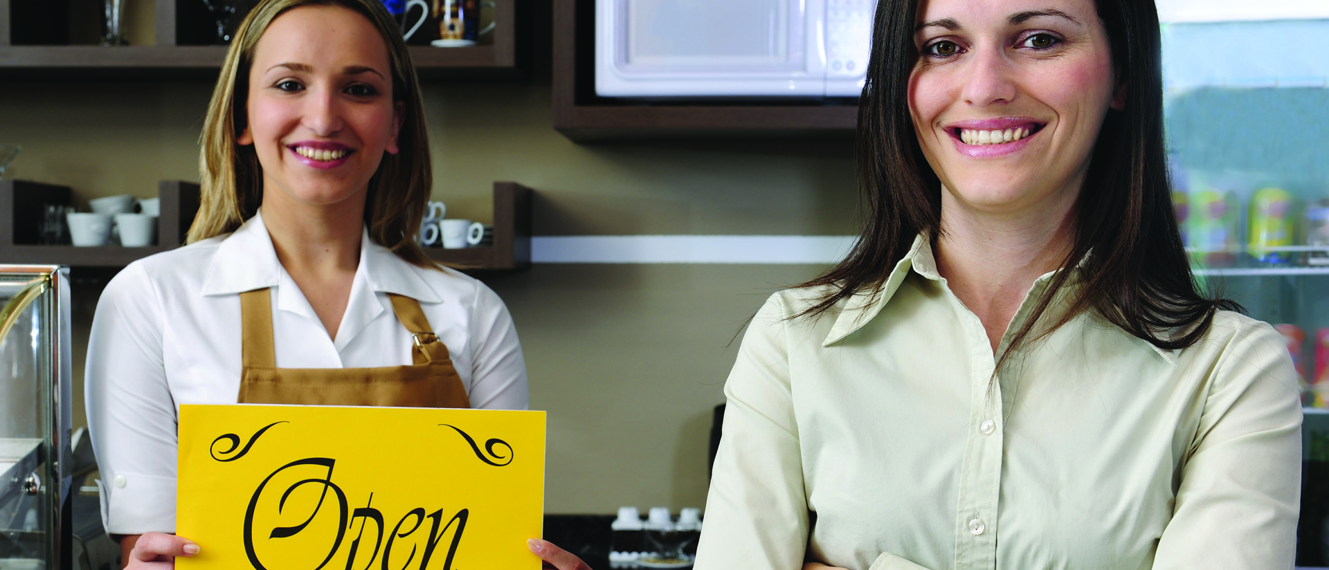Two women proudly standing in front of cafe counter with a bright yellow open sign. 