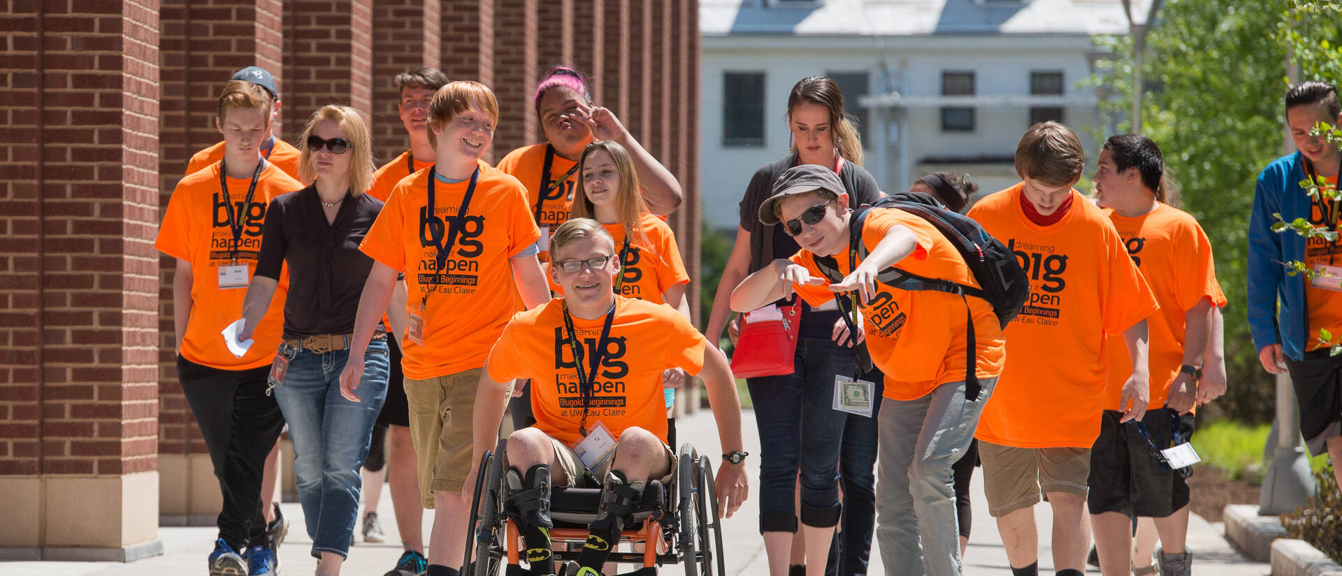 A group of students in bright orange t-shirts are smiling and messing around in front of the camera on a sunny day.